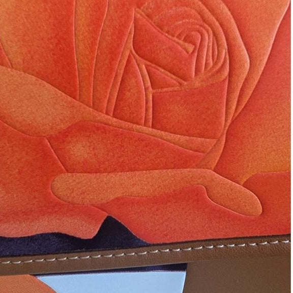 A close-up of a brown leather journal cover featuring an embossed orange rose design and a small pocket with a light blue insert. Pre-Owned Coach Tom Wesselmann Rogue Top Handle BagPre-Owned Coach Tom Wesselmann Rogue Top Handle Bag