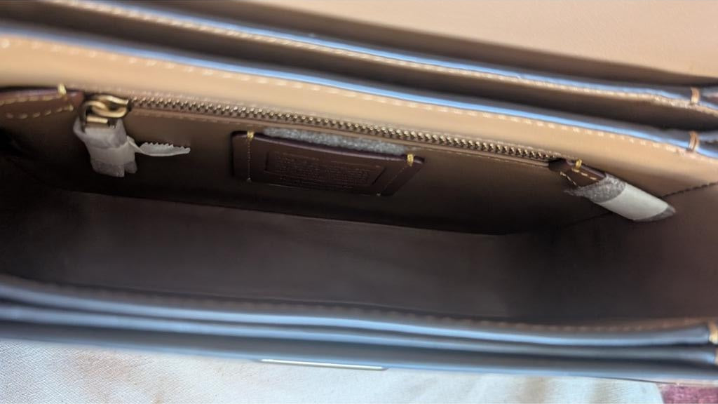 Close-up interior view of a handbag showing a beige interior lining, a dark brown zippered pocket with gold-tone hardware, and a white hangtag. Pre-Owned Coach Tom Wesselmann Rogue Top Handle Bag