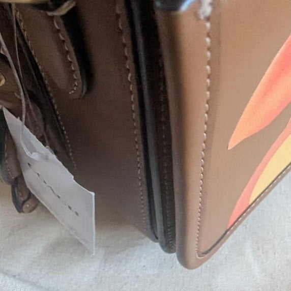 Close-up of a brown leather handbag corner showing a zip pocket, gold-tone hardware, white stitching, and a hanging product tag on a light fabric surface. Pre-Owned Coach Tom Wesselmann Rogue Top Handle Bag