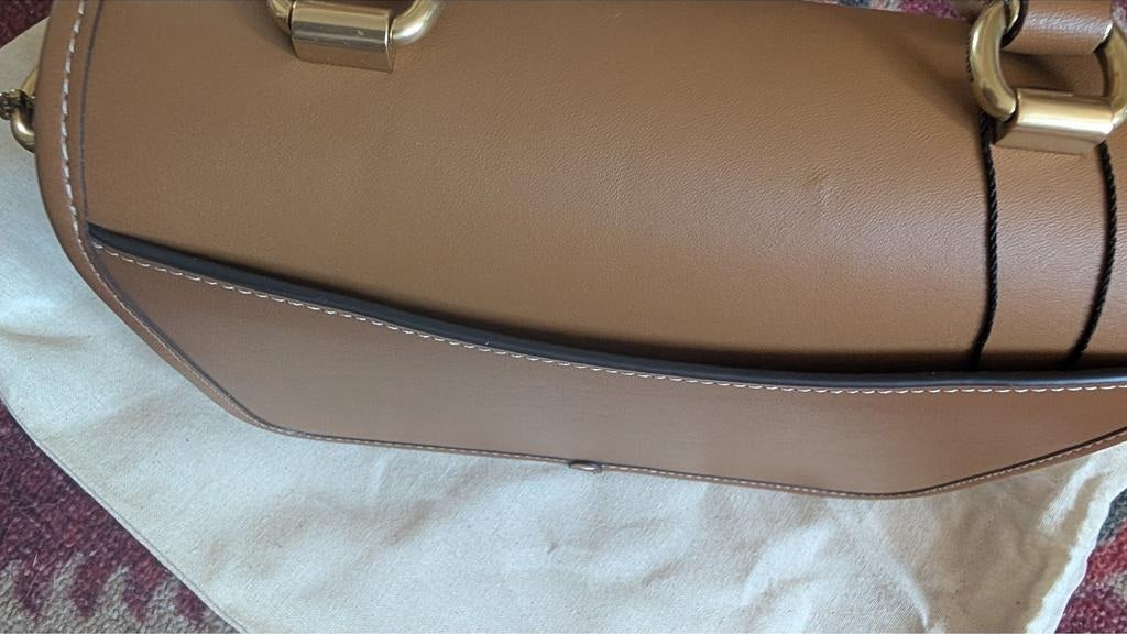 A close-up of a tan leather handbag showing the side profile, gold-tone hardware, and a black trim detail, resting on a white fabric surface. Pre-Owned Coach Tom Wesselmann Rogue Top Handle Bag