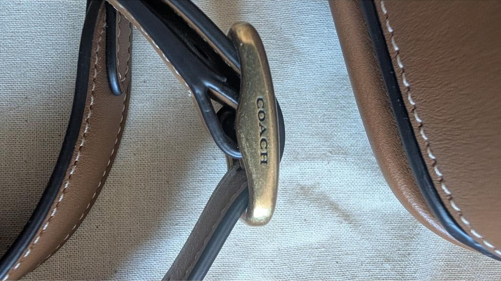 Close-up of a Coach handbag strap with a brass-tone logo clasp on a light beige canvas background. Pre-Owned Coach Tom Wesselmann Rogue Top Handle Bag