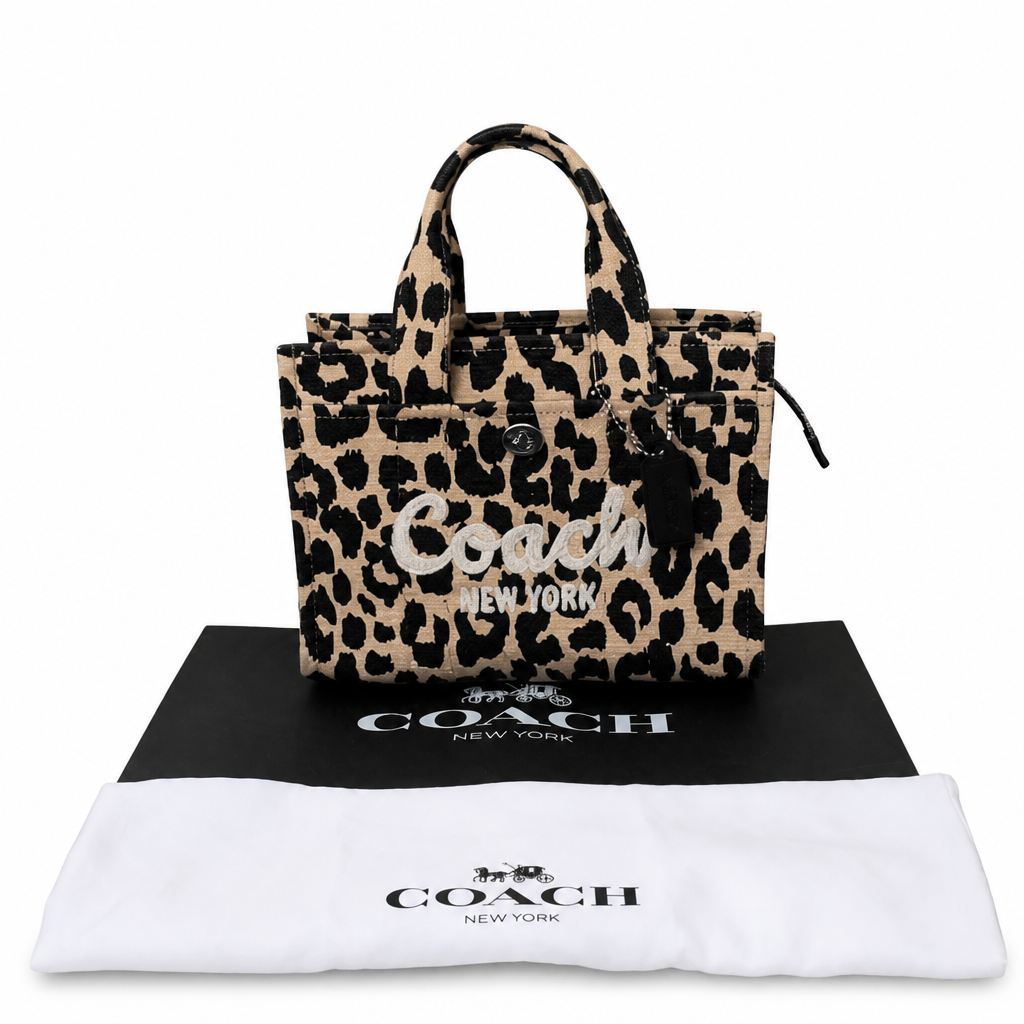 A Coach leopard print tote bag displayed on top of a black Coach dust bag with a white Coach dust bag in front, against a plain white background. Pre-Owned Coach Cargo Tote  Leopard Print Canvas Bag