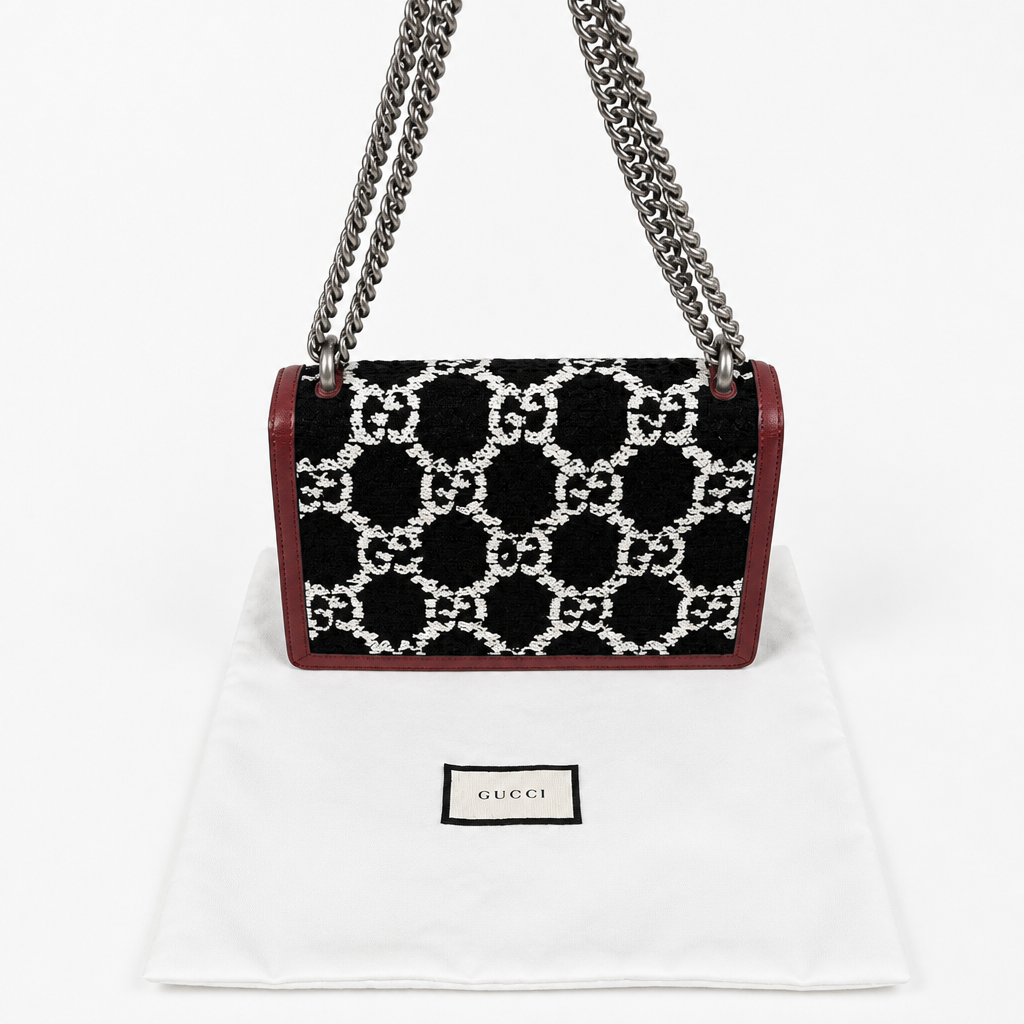 A Gucci shoulder bag with a black and white GG monogram pattern, burgundy leather trim, and silver chain straps, displayed on a white dust bag with a Gucci label. Pre-Owned Gucci Dionysus GG Tweed Medium Shoulder Bag – Red/Black