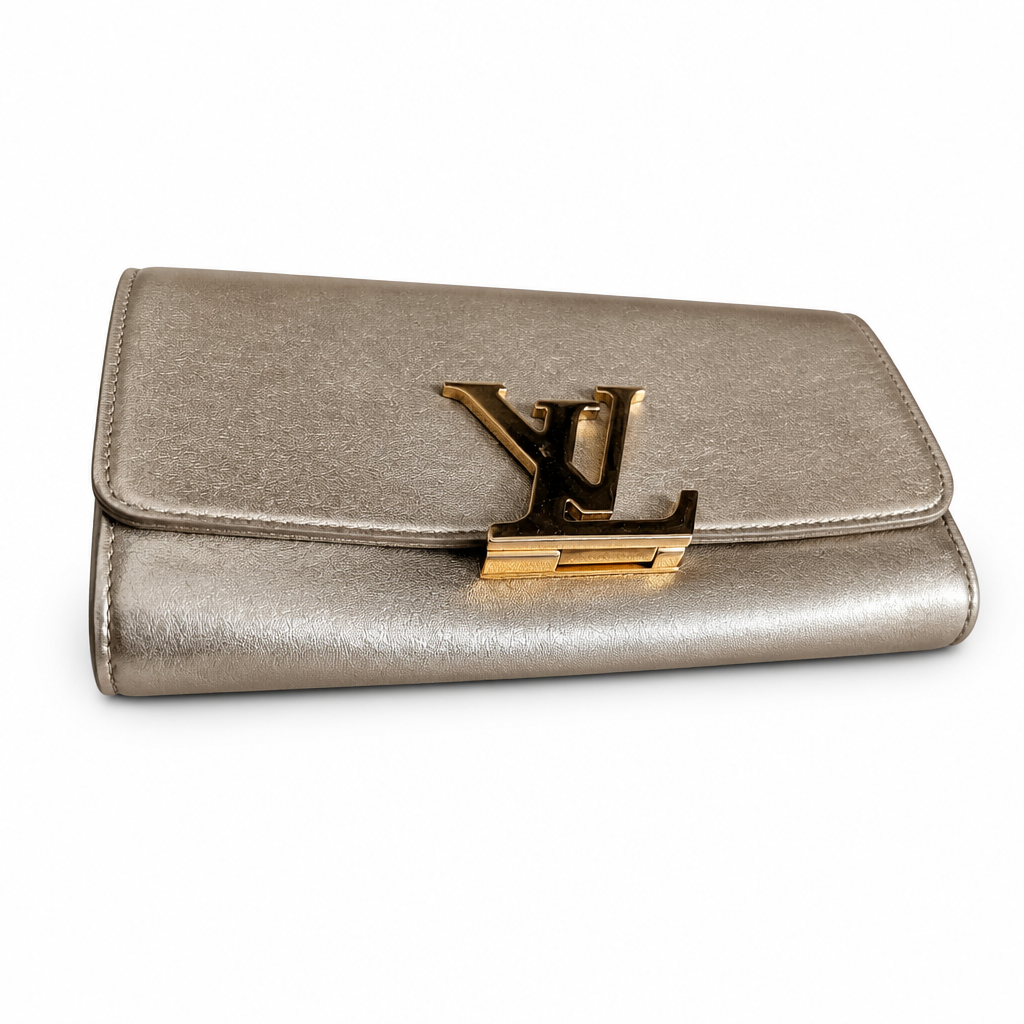 A metallic leather clutch bag with a prominent gold-tone Louis Vuitton LV logo clasp on the front flap, set against a plain white background. Pre-Owned Louis Vuitton Louise GM Clutch – Metallic Champagne Leather