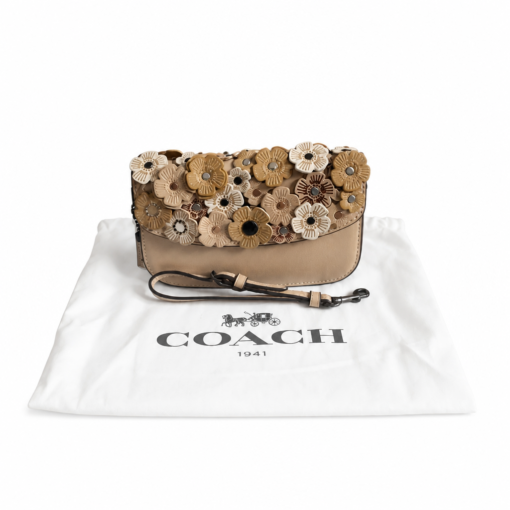 A Coach shoulder bag with a beige leather base and a top flap covered in 3D floral appliqués in tan, brown, and cream, displayed on top of its white Coach dust bag. Pre-Owned Coach Courier Wristlet with Floral Appliqué – Glovetanned Leather