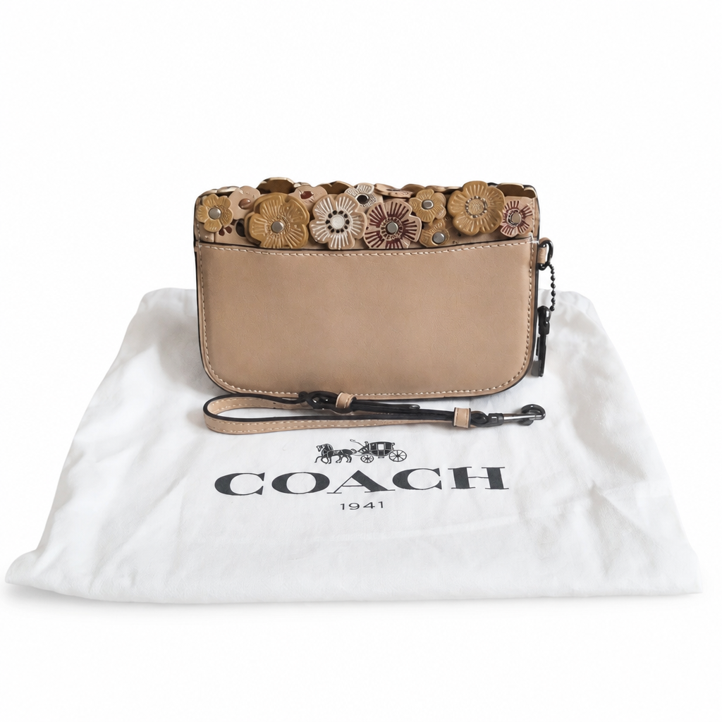 A Coach flap shoulder bag in beige leather with a floral applique trim, resting on a white dust bag printed with the Coach logo. Pre-Owned Coach Courier Wristlet with Floral Appliqué – Glovetanned Leather