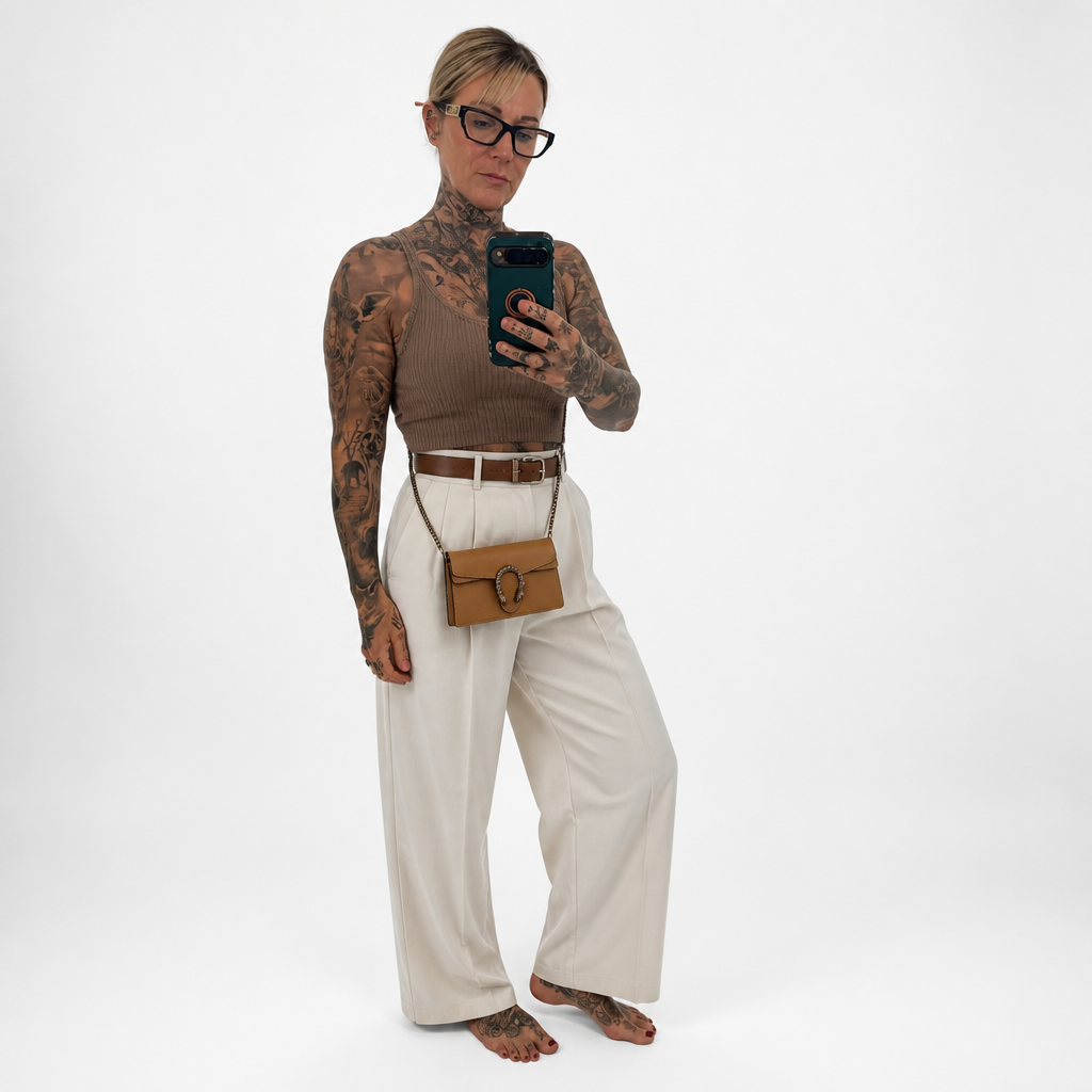 A tattooed woman takes a mirror-style selfie wearing a ribbed brown mock-neck top, cream wide-leg trousers, a brown belt, and a small brown crossbody bag with a horsebit detail. She is standing against a plain white studio background. Pre-Owned Gucci Dionysus Super Mini Crystal Tiger Head Chain Bag – Beige Leather