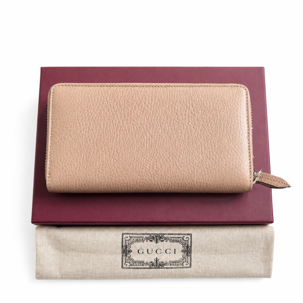 A beige pebbled leather zip-around wallet by Gucci, displayed on top of a dark burgundy box and a light beige Gucci dust bag with a visible Gucci logo patch. Pre-Owned Gucci Interlocking G Zip-Around Continental Purse in Nude/Taupe Leather