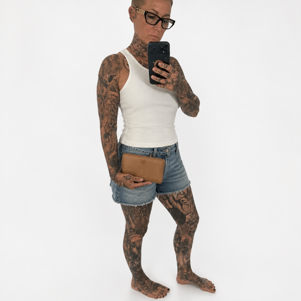 A tattooed person taking a mirror selfie wearing a white ribbed tank top, denim shorts, and glasses, holding a black smartphone and a brown leather clutch against a plain white background. Pre-Owned Gucci Interlocking G Zip-Around Continental Purse in Nude/Taupe Leather