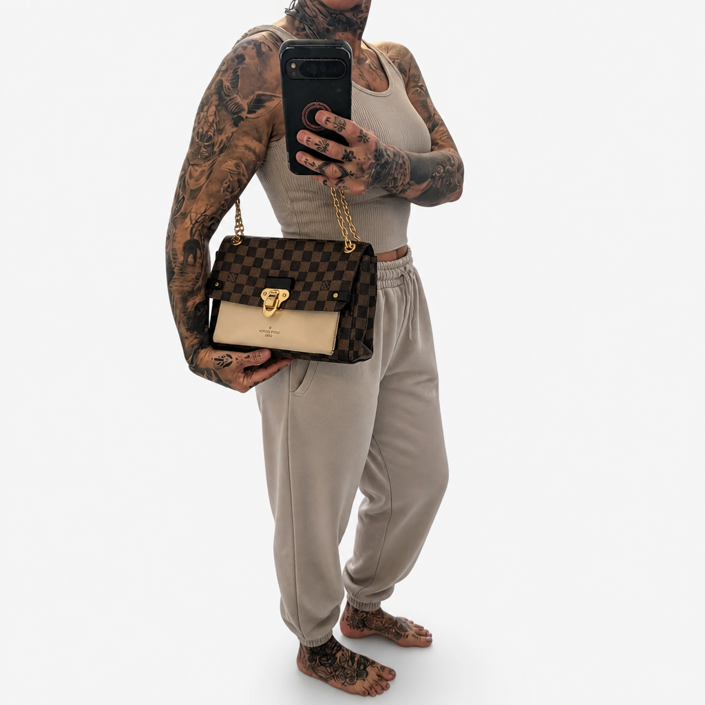 A tattooed model takes a mirror selfie while wearing a brown and beige checkered shoulder bag with a gold chain strap. The model is dressed in a beige tank top and beige sweatpants against a plain white background. Pre-Owned Louis Vuitton Vavin PM Damier Ebene / Crème – N40113