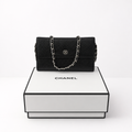 A black Chanel shoulder bag with a silver chain strap and interlocking CC logo, displayed on top of a white Chanel gift box against a clean white background. 