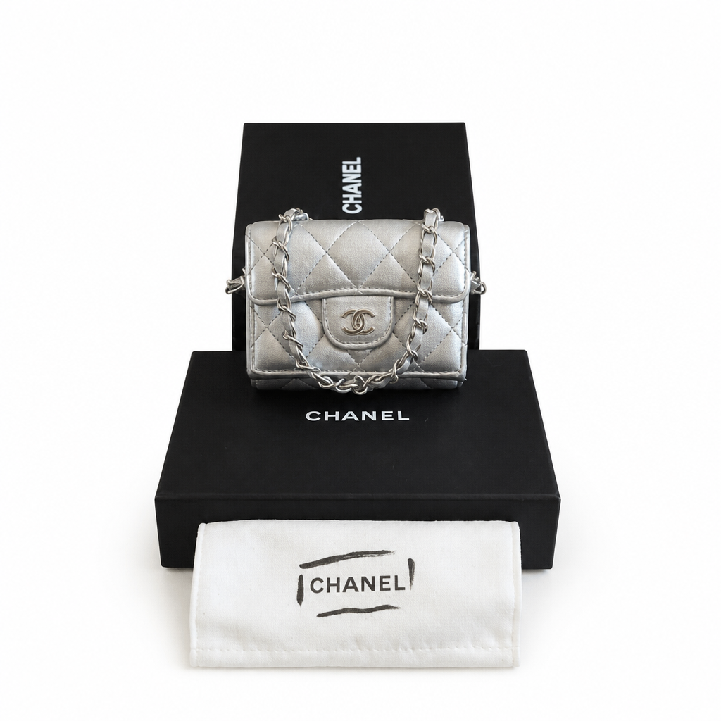A silver quilted Chanel mini shoulder bag with a chain strap, displayed on top of a black Chanel box with a white dust bag in front, against a white background. Pre-Owned Chanel Metallic Silver Matelassé Mini Chain Wallet / Micro Crossbody