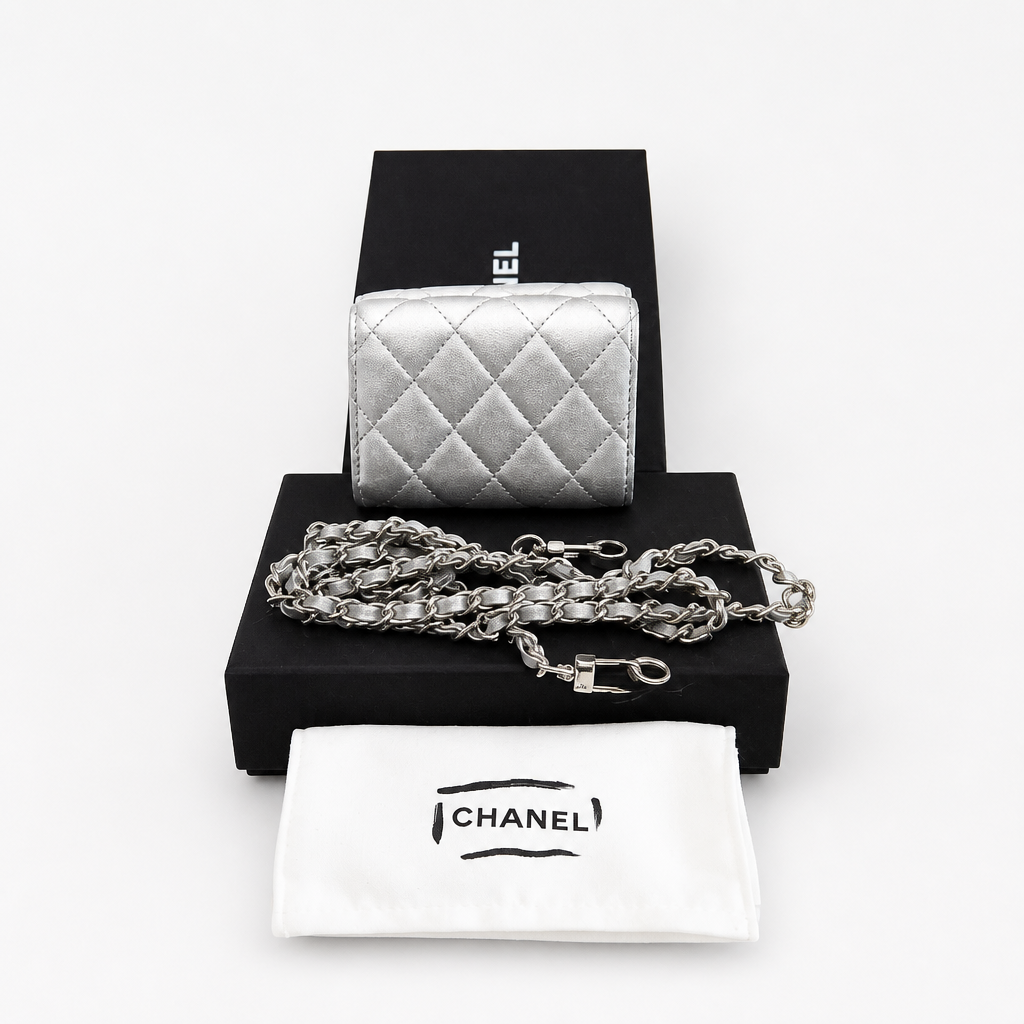 A silver quilted leather Chanel coin purse displayed on a black presentation box with its matching white dust bag and chain strap in front, on a clean white background. Pre-Owned Chanel Metallic Silver Matelassé Mini Chain Wallet / Micro Crossbody