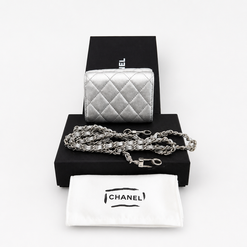 A silver quilted leather Chanel coin purse displayed on a black presentation box with its matching white dust bag and chain strap in front, on a clean white background. Pre-Owned Chanel Metallic Silver Matelassé Mini Chain Wallet / Micro Crossbody