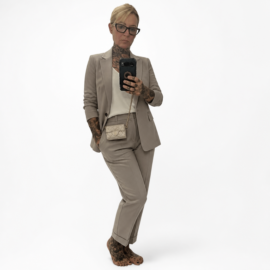 A woman with glasses and tattoos takes a mirror selfie wearing a tailored taupe suit, a white top, a small beige crossbody bag, and leopard-print boots against a plain white studio background. Pre-Owned Chanel Metallic Silver Matelassé Mini Chain Wallet / Micro Crossbody