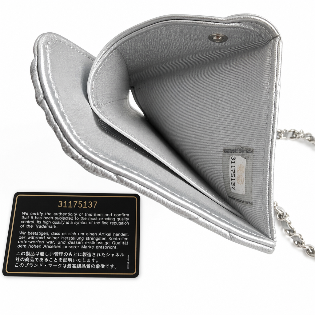 A silver metallic heart-shaped purse with a chain strap, shown open to reveal its interior compartments. A black authenticity card is placed in front of the purse on a plain white background. Pre-Owned Chanel Metallic Silver Matelassé Mini Chain Wallet / Micro Crossbody