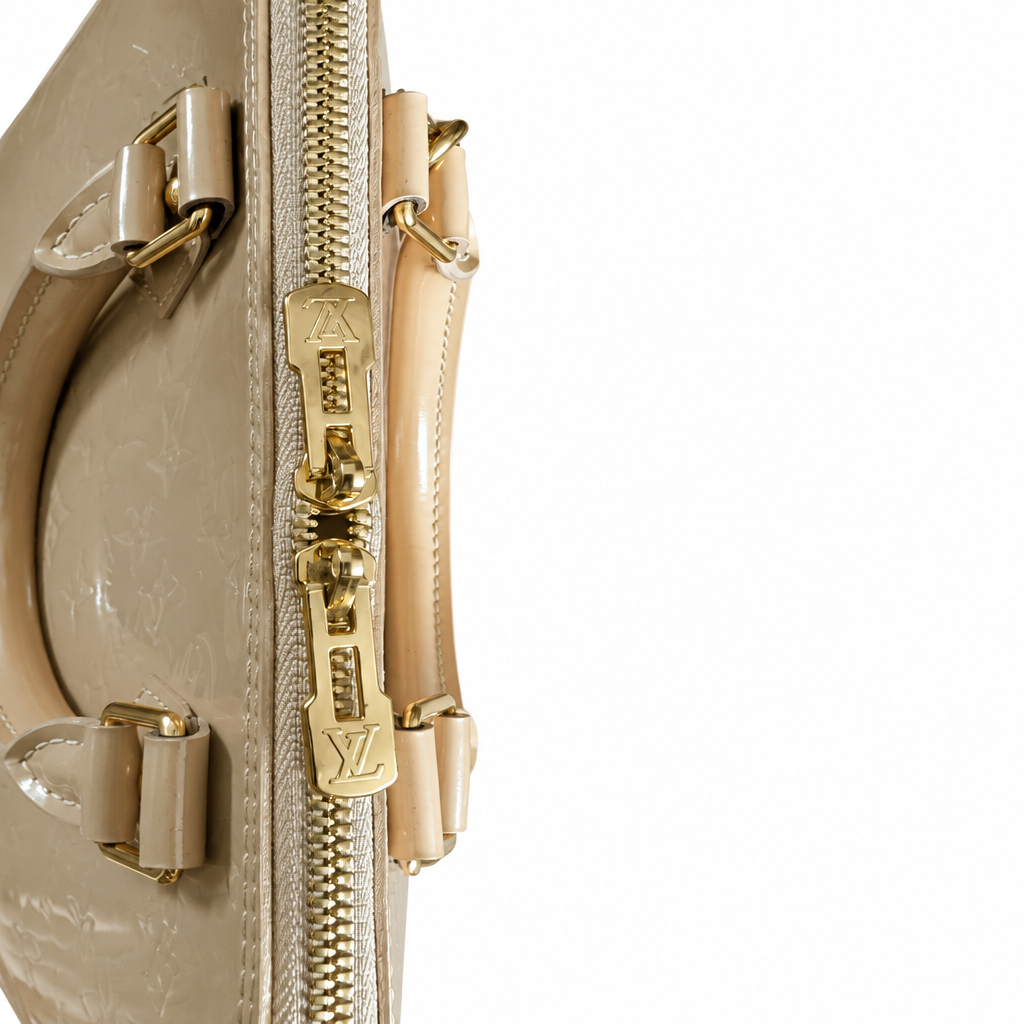 Close-up of a Louis Vuitton handbag in beige monogrammed leather with gold-tone hardware and zipper pull details, shown against a plain white background. Pre-Owned Louis Vuitton Alma PM Monogram Vernis – Beige Poudre