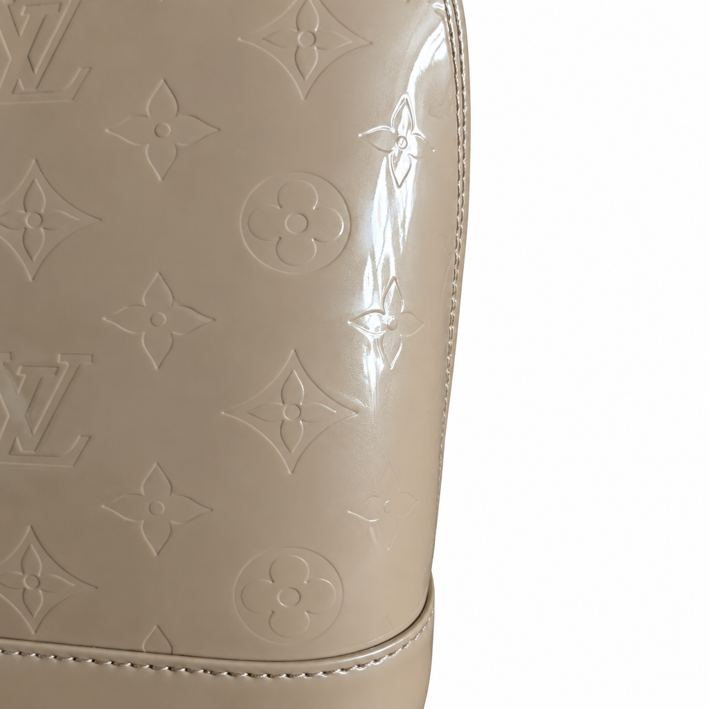 Close-up of a beige Louis Vuitton monogram bag in glossy patent leather, showing the embossed LV and floral monogram pattern against a white background. Pre-Owned Louis Vuitton Alma PM Monogram Vernis – Beige Poudre