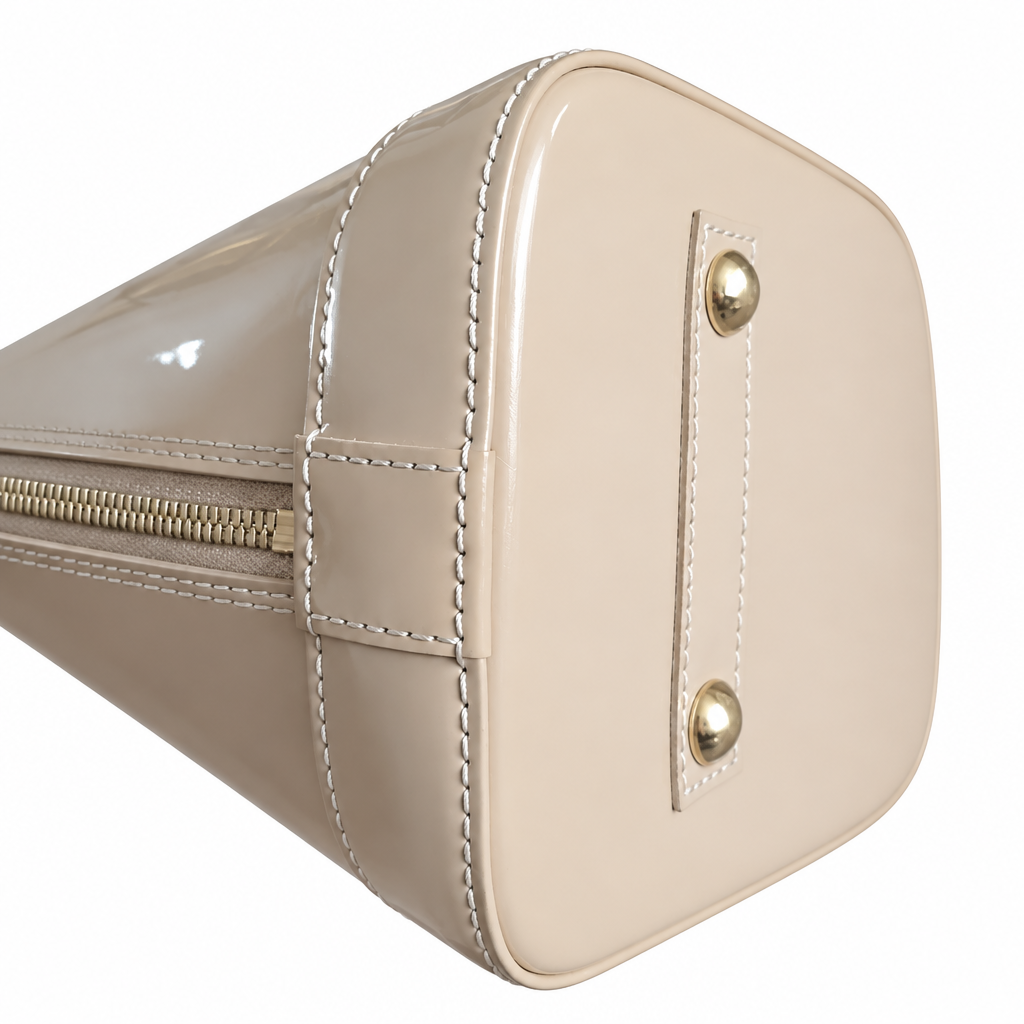Close-up of a beige patent leather handbag showing the bottom corner, white contrast stitching, a gold zipper, and gold-tone metal rivets on a white background. Pre-Owned Louis Vuitton Alma PM Monogram Vernis – Beige Poudre