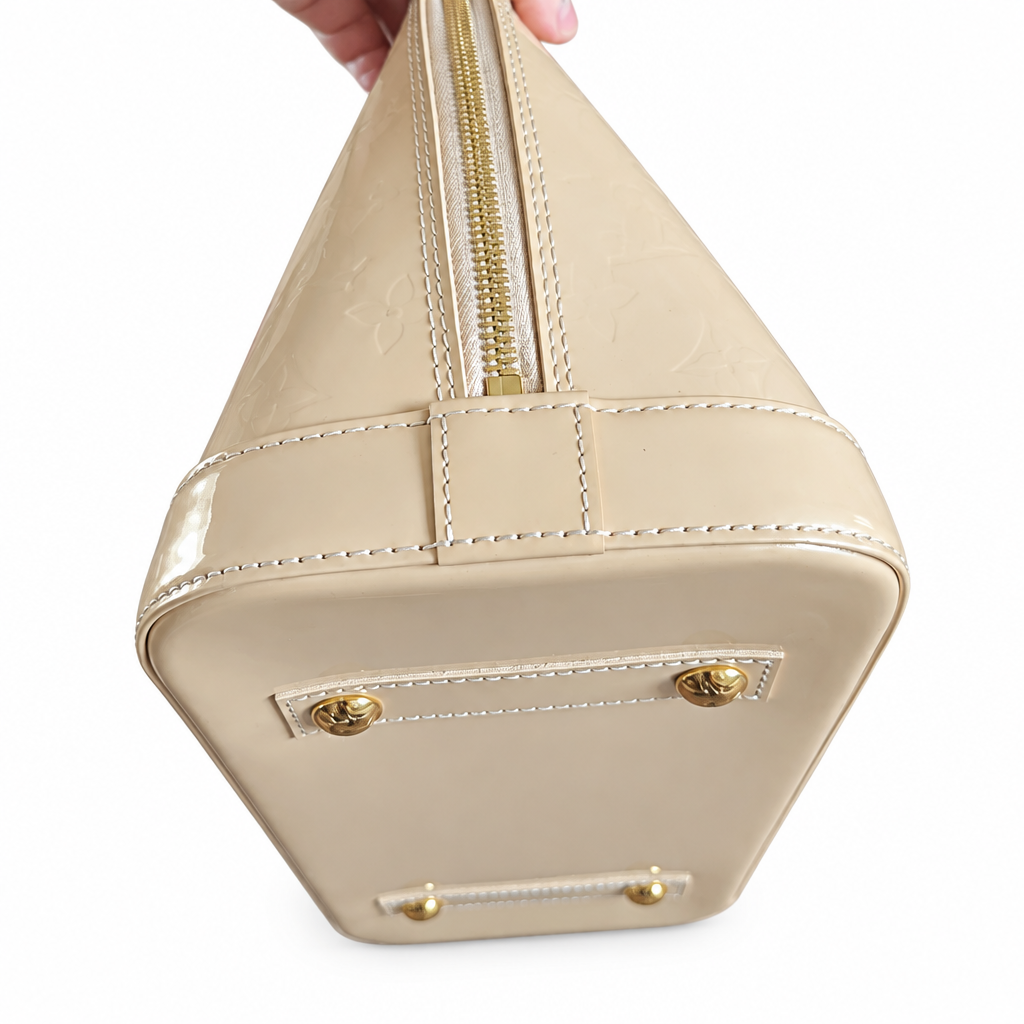 A close-up of the bottom corner of a beige patent leather handbag, showing a gold zipper, white contrast stitching, and gold-tone metal hardware against a white background. Pre-Owned Louis Vuitton Alma PM Monogram Vernis – Beige Poudre