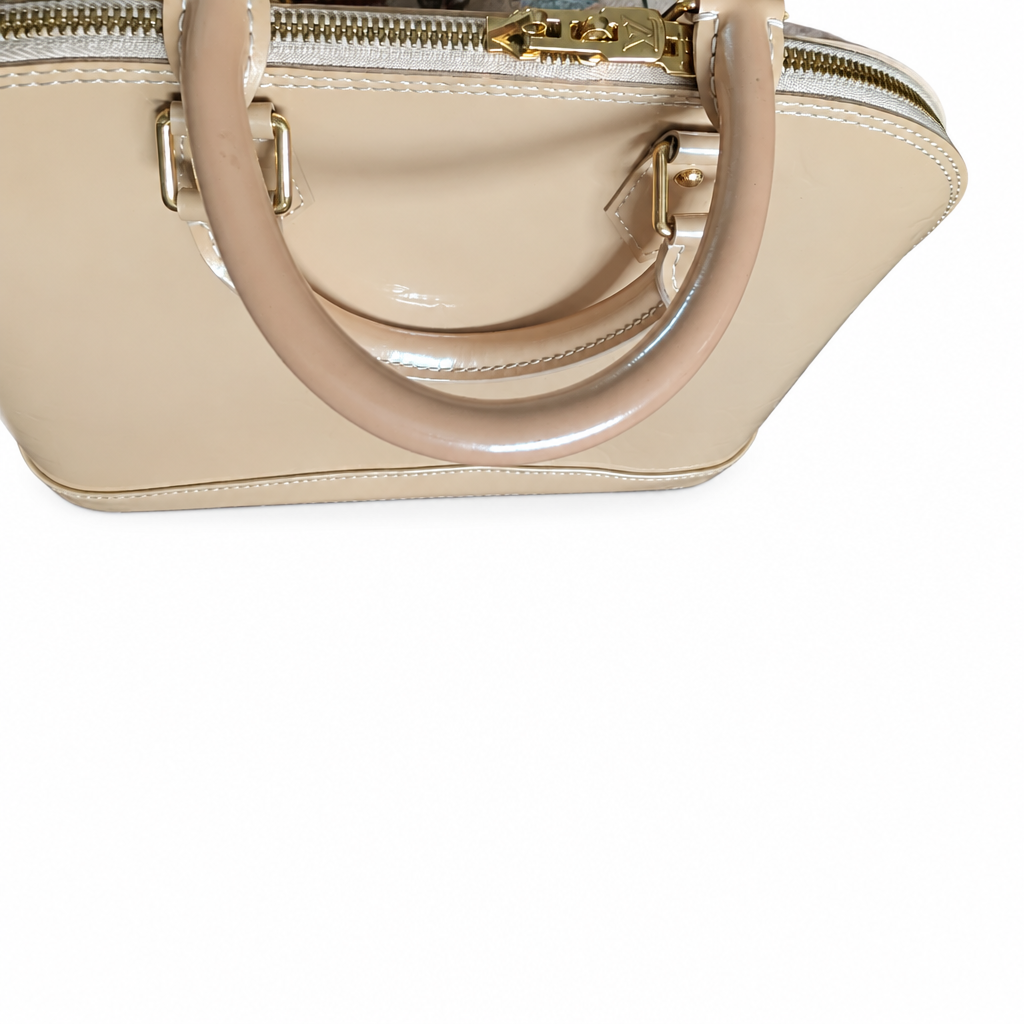 A close-up of a structured beige patent leather handbag with a glossy circular handle and gold-tone hardware, shown against a plain white background. Pre-Owned Louis Vuitton Alma PM Monogram Vernis – Beige Poudre