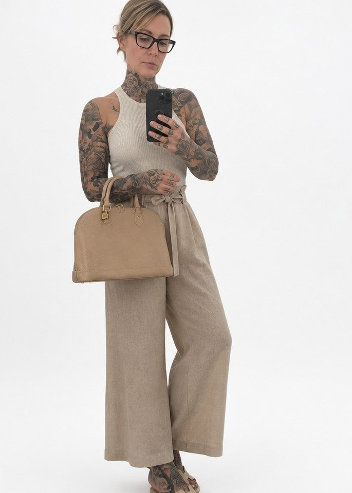 A woman with tattoos and glasses takes a mirror selfie wearing a beige sleeveless top, high-waisted wide-leg trousers, and carrying a matching beige handbag against a plain white background.  Pre-Owned Louis Vuitton Alma PM Monogram Vernis – Beige Poudre