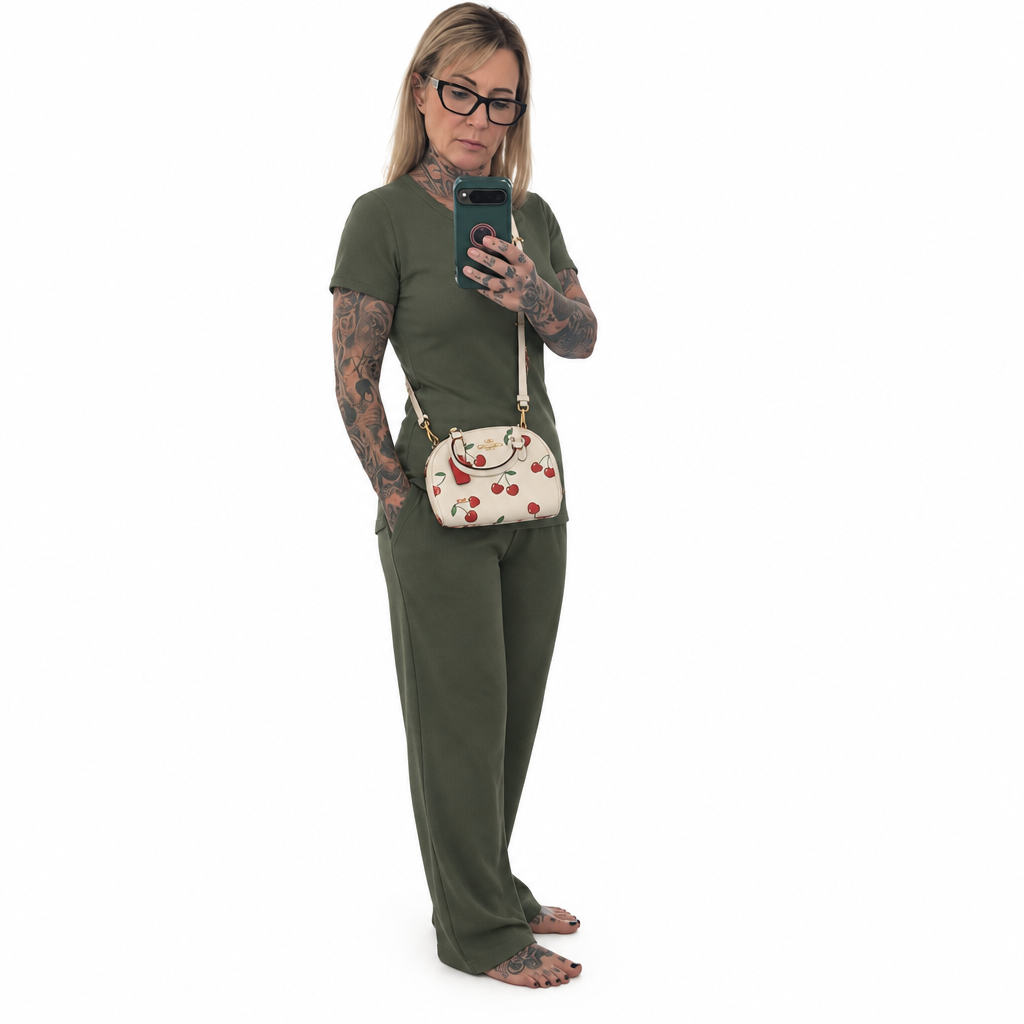 A woman modeling an olive green matching lounge set, featuring a short-sleeve crewneck top and wide-leg pants, styled with a cherry-patterned crossbody bag and glasses, set against a plain white background. Pre-Owned Coach Sydney Satchel With Heart Cherry Print – CF421