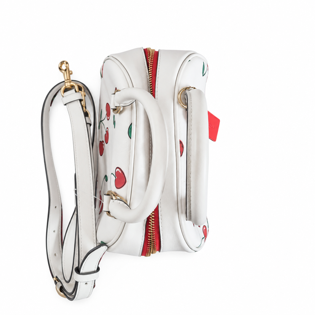 A white leather handbag with a cherry print, featuring a top handle, detachable shoulder strap, gold-tone hardware, and a red zipper, shown on a plain white background. Pre-Owned Coach Sydney Satchel With Heart Cherry Print – CF421