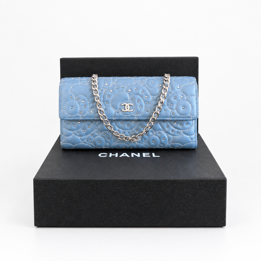 A light blue Chanel flap bag with quilted pattern, silver chain strap, and rhinestone embellishments, displayed on top of a black Chanel box. Pre-Owned Chanel Camellia Studded Wallet on Chain WOC – Blue Lambskin