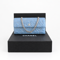 A light blue Chanel flap bag with quilted pattern, silver chain strap, and rhinestone embellishments, displayed on top of a black Chanel box. Pre-Owned Chanel Camellia Studded Wallet on Chain WOC – Blue Lambskin