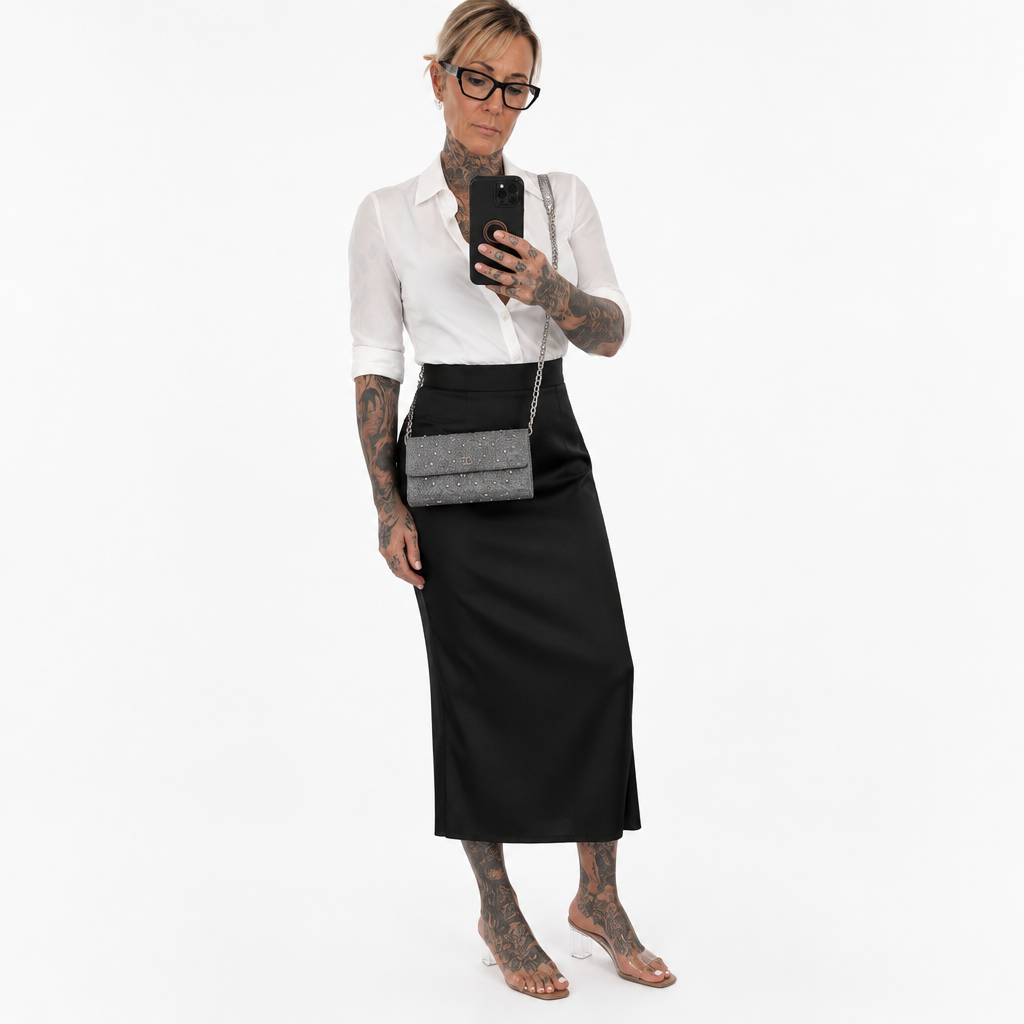 A woman modeling a white short-sleeve button-up shirt and a black midi skirt, holding a black smartphone to take a mirror selfie. She is wearing a small gray textured crossbody bag with a silver chain strap and clear high-heel sandals, set against a plain white studio background. Pre-Owned Chanel Camellia Studded Wallet on Chain WOC – Blue Lambskin