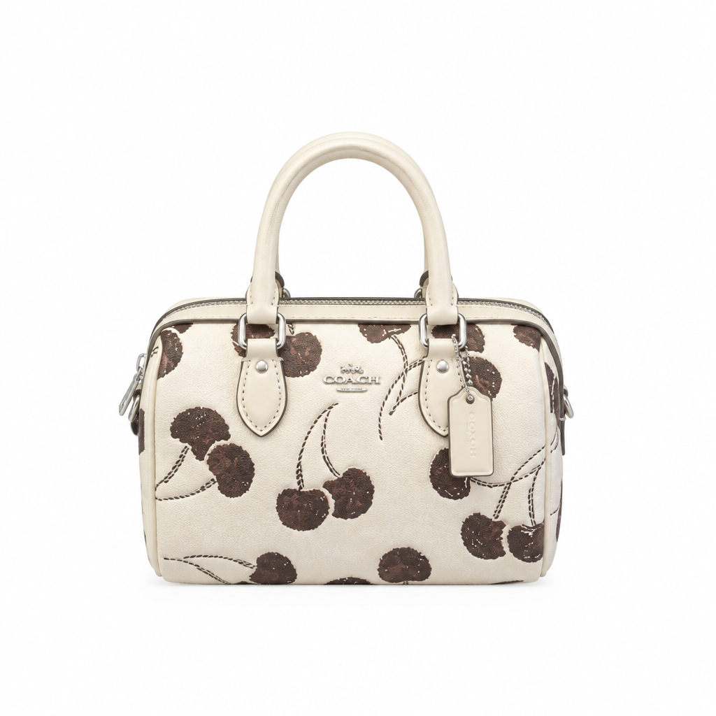 A Coach handbag featuring a cream-colored leather base with a dark cherry pattern, complete with dual top handles and a leather brand tag. Pre-Owned Coach Rowan Cherry Print Leather Crossbody / Mini Satchel – Chalk Multi