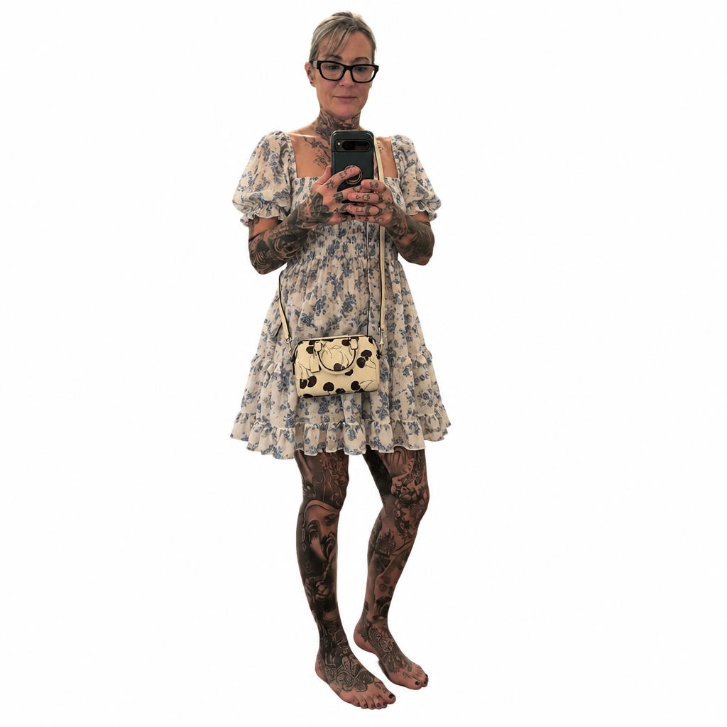 A woman with tattoos takes a mirror selfie wearing a cream-colored puff-sleeve dress with a blue floral pattern, a matching crossbody bag, and glasses against a plain white background. Pre-Owned Coach Rowan Cherry Print Leather Crossbody / Mini Satchel – Chalk Multi