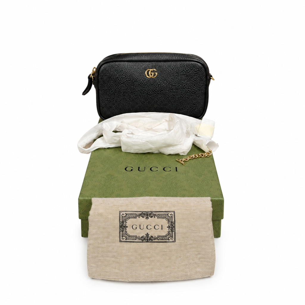 A black textured leather Gucci shoulder bag with a gold interlocking GG logo, displayed with its green Gucci box, white dust bag, and beige dust bag on a white background. Pre-Owned Gucci GG Marmont Black Leather Camera Crossbody Bag – Gold Hardware