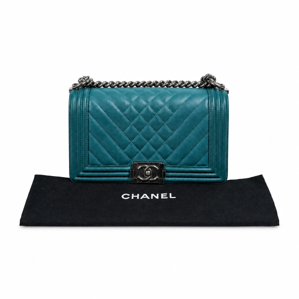 A Chanel Boy Bag in teal quilted leather with silver-tone hardware, displayed on top of a black Chanel dust bag against a white background. Pre-Owned Chanel Boy Hang Bag – Teal Quilted Leather with Ruthenium-Tone Hardware