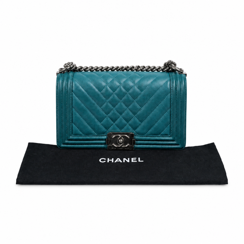 A Chanel Boy Bag in teal quilted leather with silver-tone hardware, displayed on top of a black Chanel dust bag against a white background. Pre-Owned Chanel Boy Hang Bag – Teal Quilted Leather with Ruthenium-Tone Hardware
