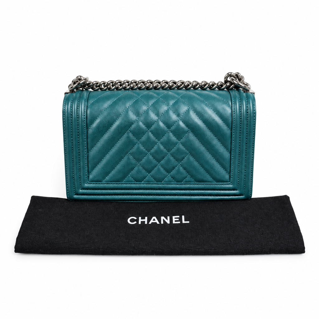 A Chanel shoulder bag in teal quilted leather with a silver chain strap, displayed above a black Chanel dust bag on a white background. Pre-Owned Chanel Boy Hang Bag – Teal Quilted Leather with Ruthenium-Tone Hardware