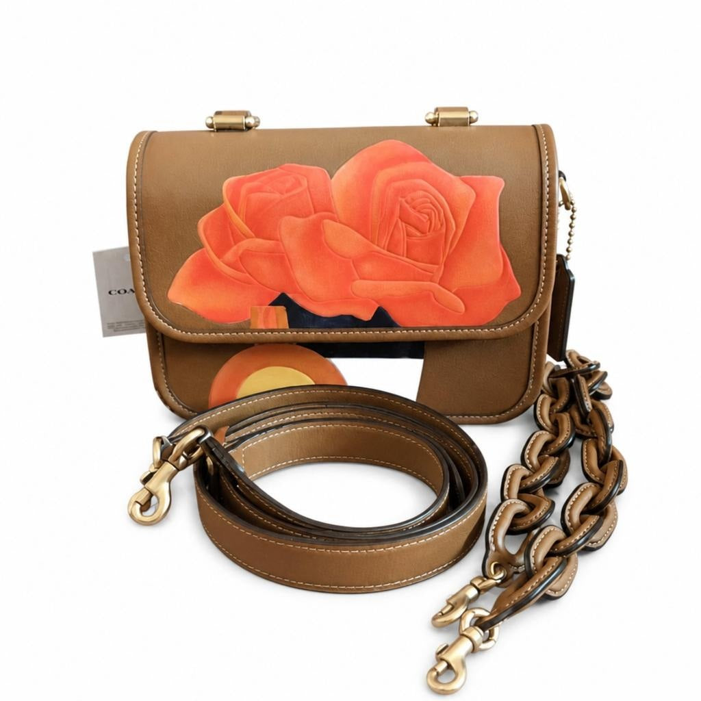 A brown leather shoulder bag featuring a large orange rose painting on the front flap, shown with its matching brown leather strap and gold-tone hardware on a white background. Pre-Owned Coach Tom Wesselmann Rogue Top Handle Bag