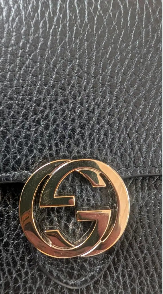 Close-up of a black textured leather item featuring a polished gold-tone interlocking G logo clasp, characteristic of a Gucci accessory. Pre-Owned Gucci Interlocking G Wallet on Chain – Black Leather
