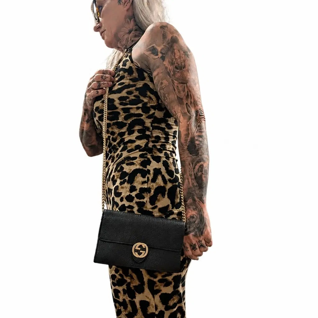 A woman with extensive tattoos on her arms and neck is shown from the side, wearing a leopard-print dress and carrying a black leather crossbody handbag with a gold-tone chain strap and interlocking G logo. Pre-Owned Gucci Interlocking G Wallet on Chain – Black Leather