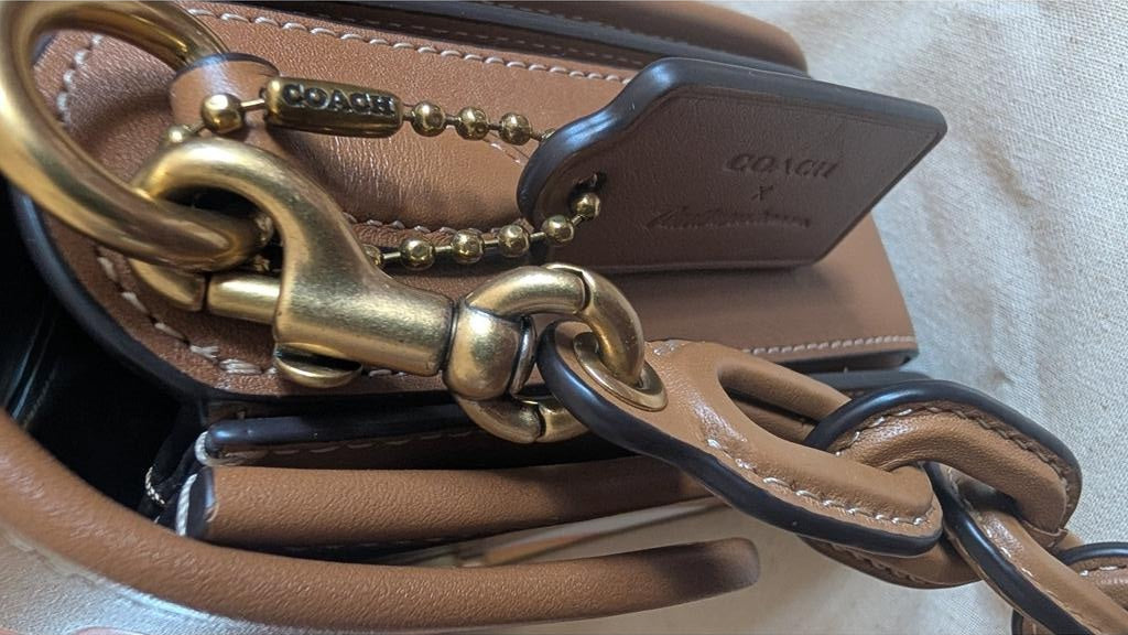 Close-up of a Coach handbag in tan leather with gold-tone hardware, a chain strap, and a leather hangtag. Pre-Owned Coach Tom Wesselmann Rogue Top Handle Bag