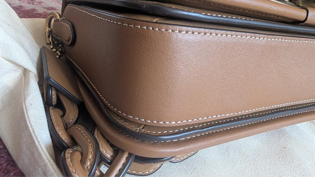 Close-up of a tan leather handbag showing the side panel, white contrast stitching, a dark trim, and part of a matching chain strap on a light fabric surface. Pre-Owned Coach Tom Wesselmann Rogue Top Handle Bag