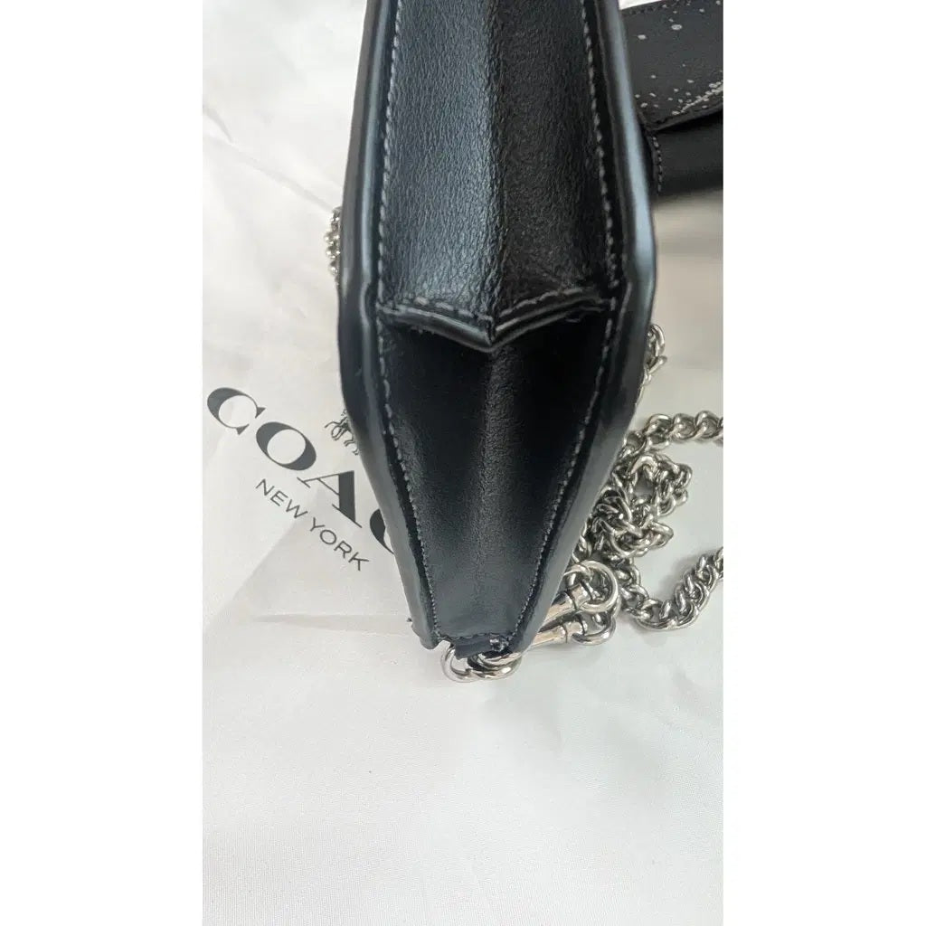 A close-up of a black leather Coach shoulder bag with a silver chain strap, resting on a white Coach New York dust bag. Pre-Owned Coach Black Celestial Print Crossbody Pouch Set – Excellent Condition