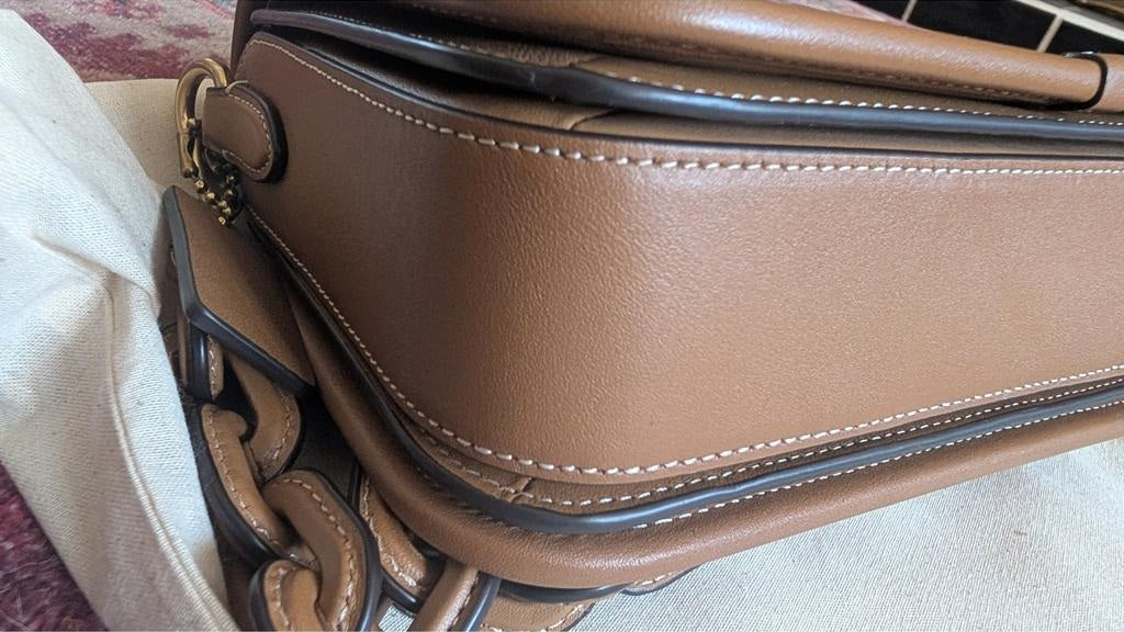 A close-up of a brown leather handbag showing the side panel, stitching, and part of a dark chain strap on a light fabric surface. Pre-Owned Coach Tom Wesselmann Rogue Top Handle Bag