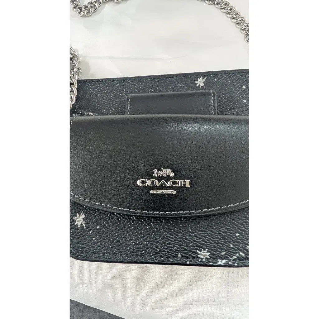 A close-up of a Coach handbag in black pebbled leather with silver-tone chain straps and a silver Coach logo on the front flap. Pre-Owned Coach Black Celestial Print Crossbody Pouch Set – Excellent Condition