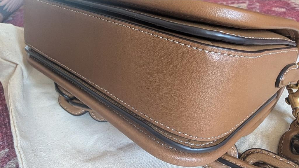 A close-up of a tan leather handbag corner showing the side panel, stitching, zipper edge, and part of a chain strap on a light fabric surface. Pre-Owned Coach Tom Wesselmann Rogue Top Handle Bag