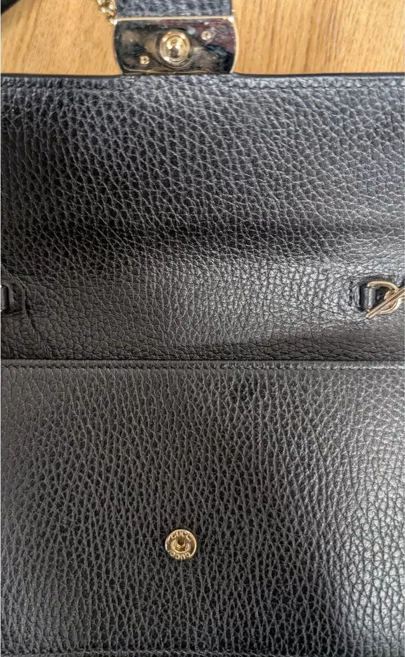 Close-up of a black pebbled leather clutch or wallet-style handbag showing the interior flap, gold-tone hardware, and part of the strap on a light wood surface. Pre-Owned Gucci Interlocking G Wallet on Chain – Black Leather
