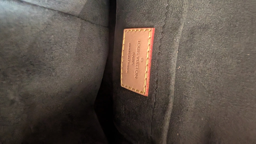 Close-up of a Louis Vuitton leather patch on a dark gray suede or velvet fabric surface. Pre-Owned Louis Vuitton Duffle Bucket Monogram Canvas - Full Set