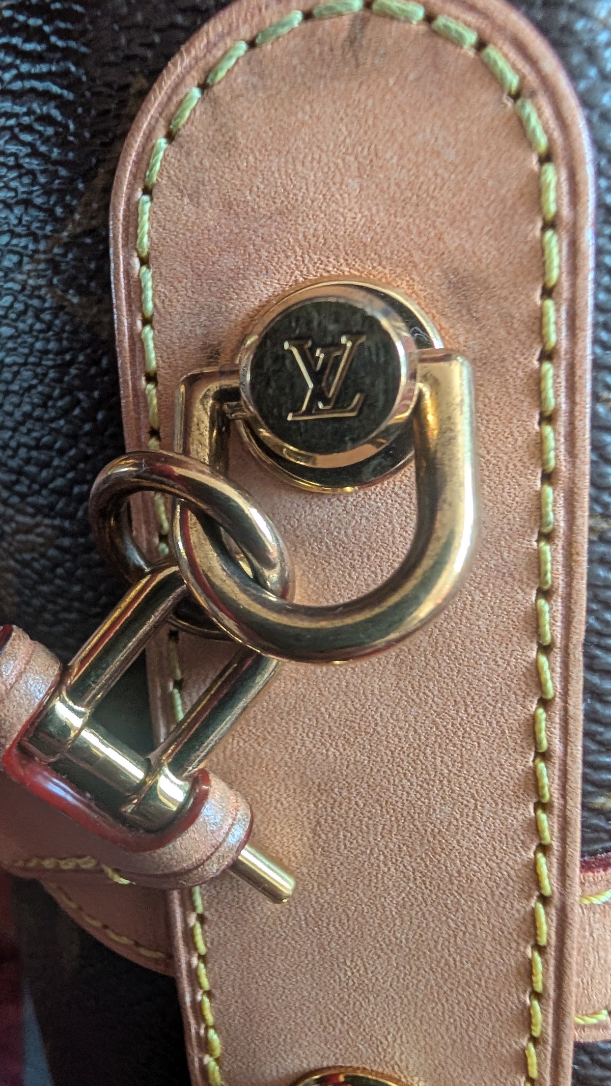 Close-up of a Louis Vuitton bag hardware detail, featuring a gold-tone metal D-ring with an engraved LV logo, a padlock clasp, and a tan leather strap on a dark monogram background. Pre-Owned Louis Vuitton Duffle Bucket Monogram Canvas - Full Set