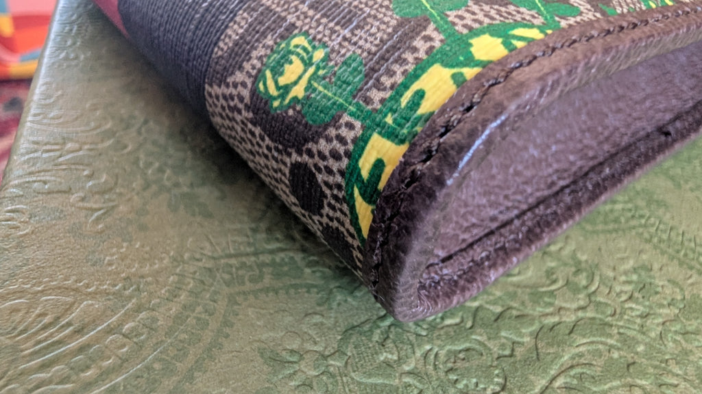 A close-up of a brown leather wallet corner with a woven fabric band featuring green, yellow, and black patterns, resting on a textured olive green surface. Pre-Owned Gucci x Ken Scott "What's Inside" Crossbody Phone Holder – GG Supreme Canvas – Full Set